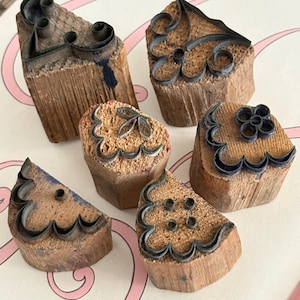 Può includere: Una collezione di sei timbri in legno vintage con vari disegni decorativi in metallo. Ogni timbro presenta un disegno in metallo scuro montato su un blocco di legno rettangolare o triangolare. I disegni includono motivi floreali e astratti, con tracce di inchiostro blu.