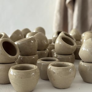 May include: A selection of small, beige ceramic pots with rounded forms and open tops. The pots are stacked, creating a textured arrangement. The neutral colour and simple design suggest a minimalist aesthetic, suitable for small plants or decorative use.