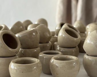 May include: A collection of small, beige ceramic pots with rounded shapes and open tops. The pots are stacked and arranged, creating a textured surface. The neutral color and simple design suggest a minimalist aesthetic, suitable for small plants or decorative use.