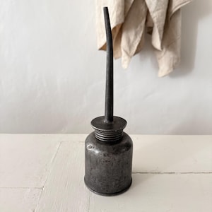 Peut inclure: Une ancienne burette d'huile en métal gris foncé avec un long bec courbé. Le bidon a un corps cylindrique et un col fileté. Le bec s'étend vers le haut, et une base circulaire est posée sur le bidon. Le bidon est posé sur une surface blanche.