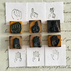 Op de afbeelding: Een set van zes vierkante houten rubberstempels, elk met een zwart handteken voor de cijfers één tot en met zes. Onder elke stempel staat een bijbehorende inkt-afdruk op wit papier die de handgebaren duidelijk weergeeft.