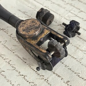 May include: A vintage metal sewing tool with a black handle and a gold-toned frame, featuring a small rotating wheel and a dark fabric ribbon. Two separate, small, circular metal attachments with intricate embossed patterns are placed nearby.
