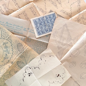May include: A collection of vintage embroidery patterns on aged paper. The patterns include floral designs, geometric shapes, and an alphabet chart in blue ink. The paper has a faded, antique appearance.