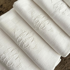 May include: A stack of white linen napkins with an embroidered leaf design. The napkins are folded and arranged in a diagonal pattern, showcasing the textured fabric and detailed embroidery. The background is a dark wood surface.