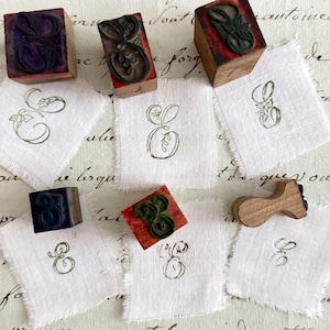 May include: An assortment of wooden letter stamps with the letter "E" and corresponding stamped images on small white fabric squares. The stamps are various colors, including purple, green, and red.