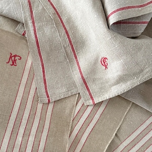 May include: Beige linen towels with red stripes and embroidered initials. The towels are folded and layered, showcasing the striped pattern and the embroidered details. The linen has a slightly textured appearance.