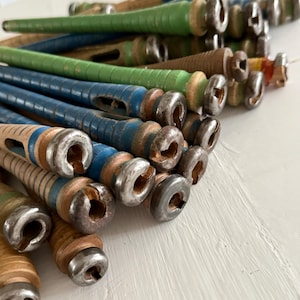 Könnte beinhalten: Eine Sammlung von Vintage-Holzspulen in verschiedenen Farben, darunter Blau, Grün und Naturholztöne. Die Spulen haben Metallenden und weisen Gebrauchsspuren auf, was darauf hindeutet, dass es sich um antike oder recycelte Artikel handelt. Die Spulen sind in einem Haufen angeordnet.