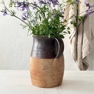 Könnte beinhalten: Ein rustikaler Keramikkrug mit zweifarbigem Design: dunkelbraun oben und hellbraun unten. Der Krug ist mit lebendigem grünen Laub und zarten lila Blüten gefüllt, was eine natürliche, ländliche Ästhetik erzeugt.