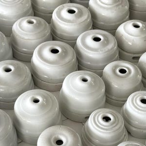 Könnte beinhalten: Eine Sammlung weißer elektrischer Isolatoren aus Keramik, die in einem Raster angeordnet sind. Jeder Isolator hat eine abgerundete, gestufte Kuppelform mit einer zentralen kreisförmigen Öffnung an der Oberseite für die elektrische Verkabelung.
