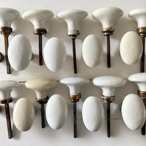 Op de afbeelding: Een verzameling antieke witte porseleinen deurknoppen met messing en ijzeren hardware. De knoppen hebben een klassieke paddenstoelvorm en ovale achterplaten. De hardware vertoont tekenen van ouderdom en slijtage.