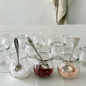 May include: A collection of small, clear glass jars with silver spoons. Some jars contain white salt, pink peppercorns, and pink salt. The jars are arranged on a white wooden surface, suggesting a kitchen or pantry setting.
