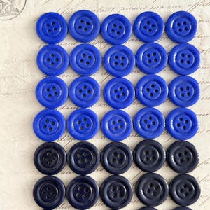 May include: A collection of round buttons in shades of blue. The buttons are arranged in rows, with a mix of bright and dark blue colors. Each button has four holes for sewing. The buttons are on a textured, off-white surface.