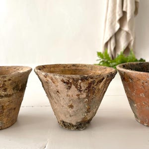 Könnte beinhalten: Drei verwitterte, konisch geformte Terrakotta-Töpfe mit strukturierten Oberflächen. Die Töpfe haben ein verblasstes, gealtertes Aussehen mit Farbtönen von Beige, Braun und einem Hauch von Grün. Die Töpfe eignen sich zum Bepflanzen von Blumen oder Kräutern.