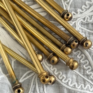 Könnte beinhalten: Eine Sammlung von Vintage-Messingstäben mit dekorativen Kugelabschlüssen und Bohrungen. Die Stäbe sind unterschiedlich lang und weisen Altersspuren auf. Der Hintergrund ist ein weißes, strukturiertes Gewebe.