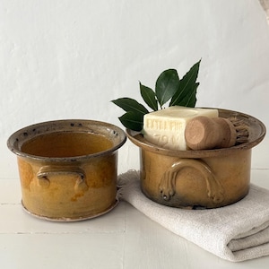 Può includere: Due vasi rotondi in ceramica smaltata con manici laterali in una calda tonalità giallo senape. Un vaso è vuoto, mentre l'altro contiene un blocco rettangolare di sapone bianco con la scritta EXTRA PUR 72%, una spazzola in legno e foglie verdi.