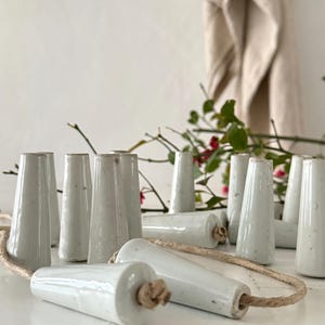 May include: A selection of white ceramic light pulls with a speckled finish. The pulls are cone-shaped and cylindrical, connected by a natural rope. Some pulls are upright, others are lying down.