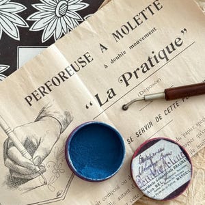 May include: An open tin of blue powder is next to a vintage document with French text. A small tool with a wooden handle and a metal tip with a spiked wheel is also visible. The document features the text "PERFOREUSE A MOLETTE" and "La Pratique".