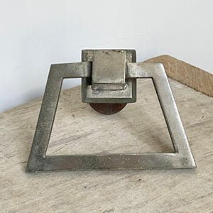 May include: A vintage metal door knocker with a geometric design. The knocker features a square central element and a triangular handle. The metal has a weathered, aged appearance, with a rusty circular base. The knocker is likely made of iron or steel.