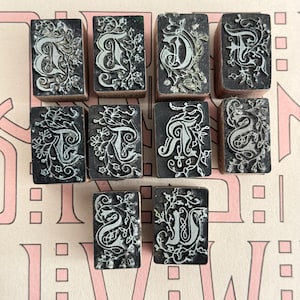 May include: A collection of ten vintage letterpress printing blocks. Each block features a different capital letter in a decorative, floral design. The letters are in a light color against a dark background. The blocks are made of wood and are rectangular.