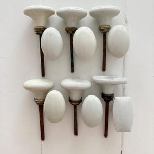 May include: A collection of white ceramic door knobs with metallic bases and spindles, including several round mushroom-shaped knobs and oval-shaped plates. The hardware features a smooth, glossy finish and shows signs of age on the metal components.