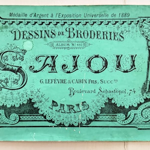 May include: A vintage green cover with black text and decorative elements. The cover reads "DESSINS DE BRODERIES" and "AJOU" in large, ornate letters. Additional text includes "Médaille d'Argent" and "PARIS."