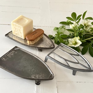 May include: Three antique-style, iron-shaped soap dishes. One holds a bar of soap and a wooden brush. The dishes are gray with a distressed finish. A sprig of white flowers and green leaves is in the background.