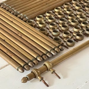 May include: A collection of vintage brass curtain rod hardware, including rods with rounded finials, mounting brackets, and screws. The brass components have a warm, aged patina, suggesting they are antique or vintage pieces. The hardware is arranged on a white surface.