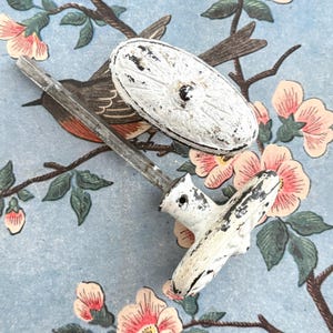 May include: Two antique white door knobs with distressed paint, one oval and one T-shaped, resting on a floral patterned background with a bird and pink flowers. The knobs have a metal rod.