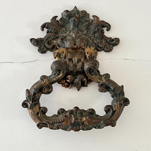 May include: Ornate bronze door knocker with a detailed floral and scroll design. The knocker has a ring handle and a decorative backplate. The metal has a distressed finish with hints of gold and green, giving it an antique appearance.