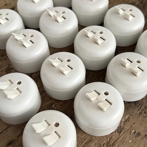 Könnte beinhalten: Eine Ansammlung weißer, runder Steckdosen mit einem Zwei-Pol-Design. Jeder Stecker hat einen weißen Schalter und ein kleines Loch. Die Stecker sind eng angeordnet und füllen den Rahmen.