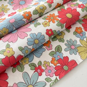 May include: A white fabric featuring a repeating floral pattern with large and small flowers in shades of red, light blue, pink, orange, and yellow, accented with green leaves. The textile has a smooth texture and is folded to show the print.