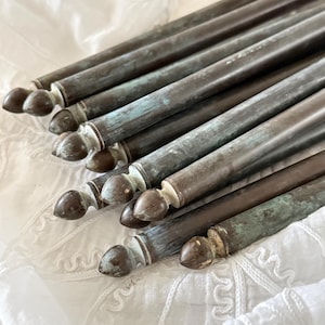 May include: A group of antique bronze curtain rods, displaying a weathered patina. Each rod features a cylindrical body and a decorative finial. The rods are arranged on a white, textured surface, highlighting their aged appearance and unique design.