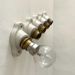 Könnte beinhalten: Reihe von Vintage-Leuchten aus weißem Porzellan mit Messing-Lampenfassungen, an einer weißen Wand befestigt. Eine klare Glühbirne aus Glas ist in eine Fassung geschraubt. Die Leuchten sind abgewinkelt.