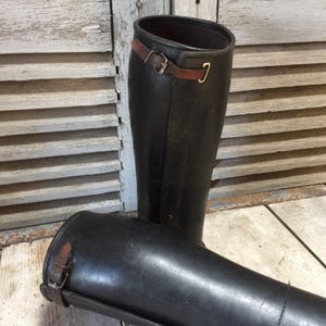 May include: A pair of tall, black leather riding boots. Each boot features a brown leather strap with a buckle near the top and a gold-colored hook. The boots are displayed on a wooden surface.