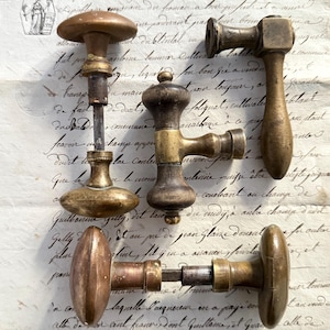Peut inclure: Un assortiment de boutons et poignées de porte anciens en laiton, de formes et tailles variées. La quincaillerie présente un aspect vieilli et patiné, avec des signes d'usure visibles. Les boutons et poignées sont présentés sur un fond de papier vieilli avec une écriture manuscrite.