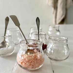 Peut inclure: Plusieurs petits pots en verre transparent avec des cuillères en argent. Certains pots contiennent du sel rose, d'autres sont vides ou remplis d'épices. Les pots sont disposés sur une surface blanche, créant une esthétique rustique et champêtre.