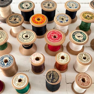 Puede incluir: Una colección de carretes de hilo de madera vintage en varios colores, incluyendo negro, rojo, blanco y verde. Los carretes tienen etiquetas con texto y números, dispuestos sobre una superficie blanca.
