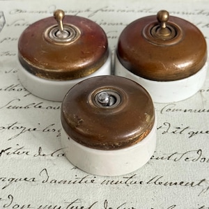 Puede incluir: Tres interruptores de luz de estilo vintage con acabado metálico marrón y bases de cerámica blanca. Cada interruptor tiene una palanca y un diseño circular, mostrando una estética antigua. Los interruptores están dispuestos sobre una superficie con escritura manuscrita.