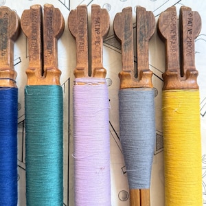 May include: A close-up of five vintage wooden thread winders, each with a different color of thread: blue, teal, lavender, gray, and yellow. The winders have a distinctive shape with a handle and a slot for the thread. The text "MASCHINENFABRIK RUTH" and "PAT.NO. 210076" is visible.