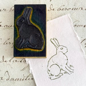 May include: A rectangular wooden rubber stamp featuring a carved black rabbit silhouette with a yellow border. Beside the stamp is a piece of textured white paper displaying a stamped green ink impression of the same rabbit in a seated position.