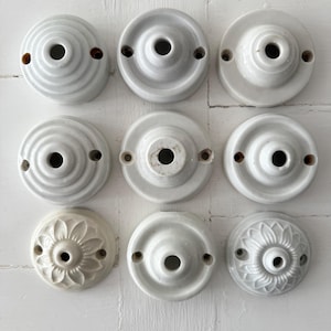 May include: Nine white ceramic ceiling rose covers with different designs. Each cover has a central hole and two mounting holes. Some have concentric rings, others feature floral patterns. Ideal for lighting.