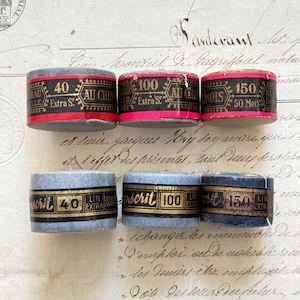 Könnte beinhalten: Sechs Vintage-Garnspulen mit Etiketten. Die Spulen sind in Grau-, Blau- und Rottönen gehalten und tragen schwarz-goldene Etiketten mit Angaben zu Garnart und -länge. Die Etiketten tragen Texte wie "40", "100" und "150".