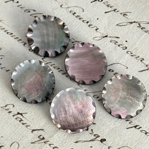 May include: Five circular buttons with a scalloped edge and a smooth, iridescent surface. Each button displays a unique mother-of-pearl finish with shifting tones of grey, soft pink, and silver, reflecting light across their polished, rounded surfaces.