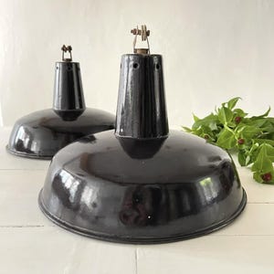 Puede incluir: Dos lámparas colgantes industriales negras vintage. Cada lámpara tiene una pantalla ancha y redondeada y una parte superior cilíndrica con un lazo de metal para colgar. El acabado negro brillante refleja la luz.