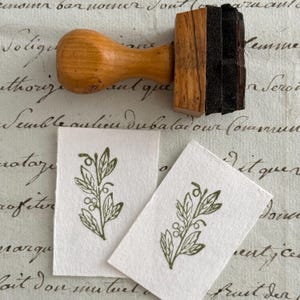 Op de afbeelding: Een houten stempel met een lichtbruine handgreep en een donkerbruin stempelkussen. Twee kleine witte kaarten tonen een groen botanisch ontwerp. De kaarten zijn gestempeld met een blad- en bessenmotief.