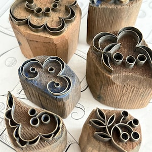 Könnte beinhalten: Eine Sammlung von sechs Holzstempeln mit Metalldesign. Die Stempel zeigen verschiedene florale und geometrische Muster, darunter Kreise, Wirbel und Blattmotive. Das Holz hat ein natürliches, gealtertes Aussehen, die Metallmuster sind silberfarben.