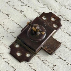 Può includere: Un fermo per finestra in metallo marrone scuro con una manopola rotante centrale con finitura in ottone usurato. La piastra di base rettangolare include due flange di montaggio con tre fori circolari per viti su ciascuna estremità e un chiavistello.