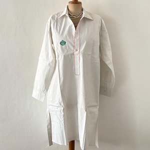 Puede incluir: Túnica blanca de manga larga con cuello y ribete rojo a lo largo de la tapeta y el cuello. Un pequeño emblema está en el pecho. La túnica se exhibe en un maniquí.