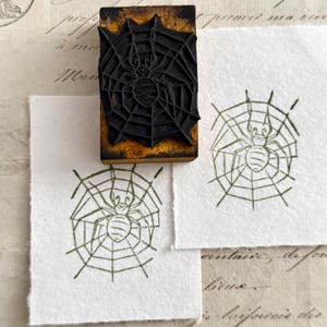 May include: A black rubber stamp with a spider and web design. The stamp is on a wooden block. Two impressions of the stamp are on white paper. The spider web design is in a circular shape.