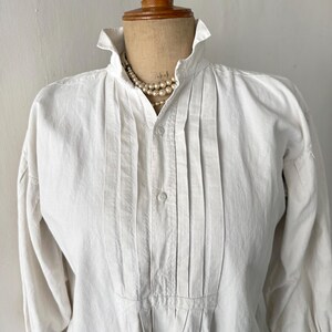 May include: A white, long-sleeved shirt with a high collar and vertical pleats down the front. The shirt has a button-down closure and is displayed on a mannequin. A pearl necklace is draped around the collar.